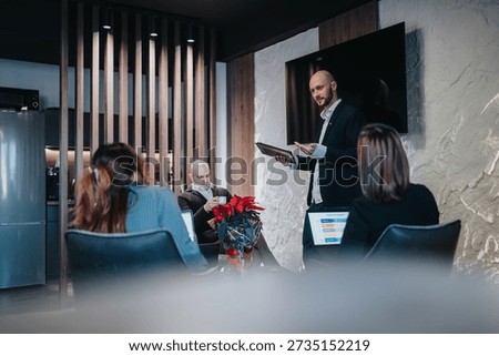 A presenter stands with tablet addressing a small group in a contemporary workspace. Team members sit with laptops and drinks, while a poinsettia adds color to the scene.