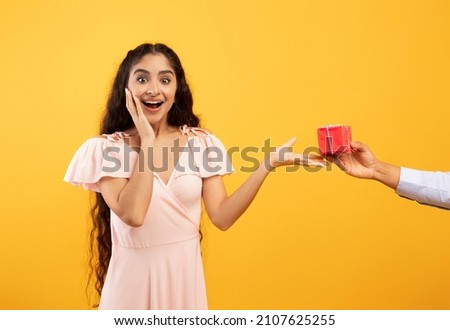 Presents delivery, holiday celebration concept. Hand holding and giving gift box to surprised excited indian woman standing over yellow studio background