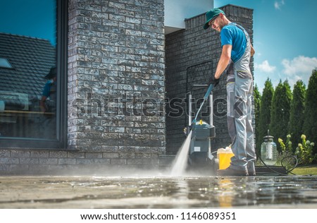 Pressure Washer Cleaning in Front of the House. Caucasian Men in His 30s Washing Concrete Bricks Driveway in Sunny Summer Day. Cleaning Around the House Concept.