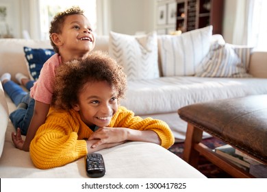 Pre-teen African American girl lying on sofa watching TV in the living room with her younger brother sitting on her back, close up - Powered by Shutterstock