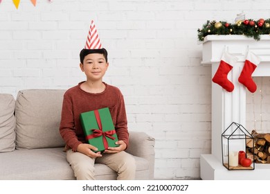 Preteen Asian Boy In Party Cap Holding Present On Couch Near Christmas Decor At Home