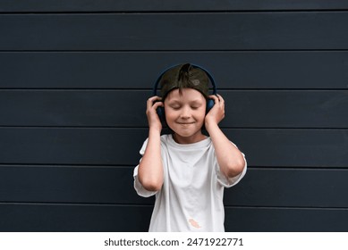 Pre-teen boy in a cap with headphones listening to music with closed eyes against a background of wooden planks in graphite color - Powered by Shutterstock