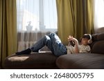  preteen boy wearing headphones listens to music and watches a video sitting on the couch at home. favorite music. Generation Z. 