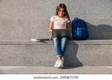 Preteen caucasian elementary middle school girl with backpack sitting on the stairs steps outside and using working laptop looking on screen doing homework project while break recess - Powered by Shutterstock