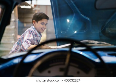 Preteen Child Looking At The Engine Of A Vintage Car From The 60s. He Is Amazed And Excited. The Boy Smiles Happy And Leans Against The Open Hood. Viewed From The Interior Of The Car.