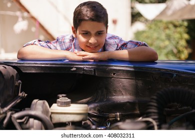 Preteen Child Looking At The Engine Of A Vintage Car From The 60s. He Is Amazed And Excited. The Boy Smiles Happy And Leans Against The Open Hood With Chin On Hands.