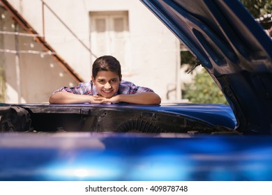 Preteen Child Looking At The Engine Of A Vintage Car From The 60s. He Is Amazed And Excited. The Boy Smiles Happy And Leans Against The Open Hood With Chin On Hands.