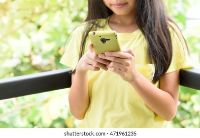 Preteen Girl With Black Hair Stand On Terrace With Mobile Phone At Home. Young Asian Student Reading Text Messages On Cell Phone. Kid Holding And Typing Message In Smart Phone. Focus On Hand.