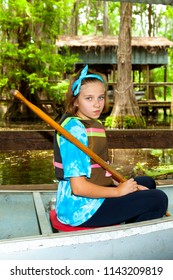 A Pre-teen Girl Sits In A Metal Canoe, Ready To Launch, With Paddle In Hand And A Smirk On Her Face.  She Is In Front Of A Swampy Background With Cypress Trees And Spanish Moss Hanging Down.