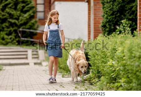Preteen girl wearing jeans dress with golden retriever dog walking outdoors in summertime. Pretty kid petting fluffy doggy pet in city