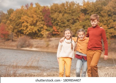 Preteen Kids, Boy And Twin Girls Walking In Fall Park With Lake.  Friends Wearing Warm Knit Sweaters