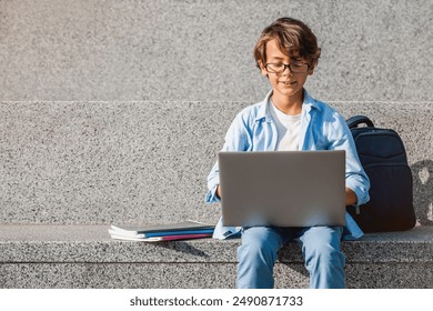 Preteen school boy in eyeglasses with backpack sitting on the stairs steps outside and working with laptop looking on screen using gadget doing homework project while break recess, copy space - Powered by Shutterstock