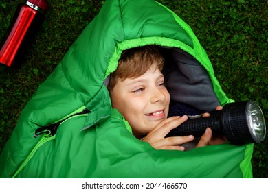 Preteen School Kid Boy In Sleeping Bag Camping. Outdoors Activity With Children In Summer. Fun And Adventure Camp, Family And Friends Vacations Or Weekend Trip. Portrait Of Child With Flashlight.