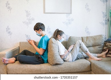 Pre-teen sister and brother gaming online with cellphones while sitting at home, children wearing safety masks, sitting back to back on sofa