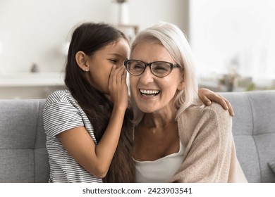 Pretty adorable granddaughter telling secret to happy laughing grandma, whispering at ear. Cheerful grandmother and preteen girl enjoying family communication, listening to kid - Powered by Shutterstock