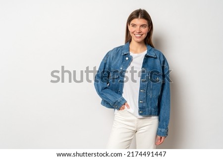 Pretty adorable youthful woman with fair hair dressed in white t-shirt, denim jacket and jeans, holding hand in pocket, smiling against blank studio wall with copy space. Fashion and trends concept