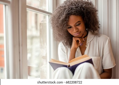 Pretty african american girl with reading a book sitting on the windowsill.