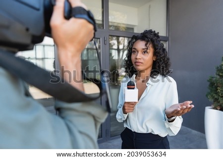 pretty african american journalist with microphone gesturing while doing reportage near blurred cameraman