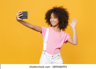 Pretty African American Kid Girl 12-13 Years Old In Pink T-shirt Doing Selfie Shot On Mobile Phone, Greeting With Hand Isolated On Yellow Background. Childhood Lifestyle Concept. Mock Up Copy Space.