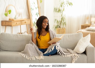 Pretty African American Woman Holding Mug Of Fresh Aromatic Coffee And Reading Interesting Book While Sitting Under Warm Plaid On Sofa At Home. Peaceful Weekend Activities Concept