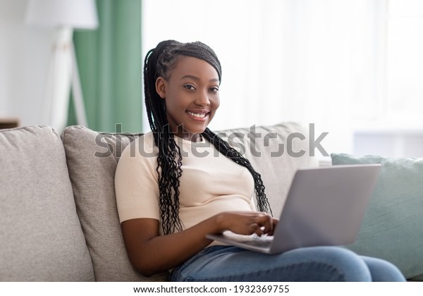 Pretty african american young lady with braids\
sitting on couch at home, using laptop, watching videos online,\
chatting with friends or reading blogs, copy space. Lifestyle\
during covid-19 pandemic