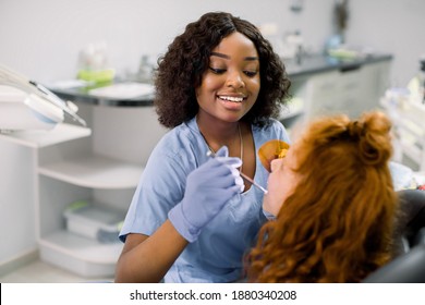 Pretty african female dentist in blue uniform and gloves, making tooth restoration and filling with curing polymerization UV lamp for her little patient, cute girl with red curly hair - Powered by Shutterstock