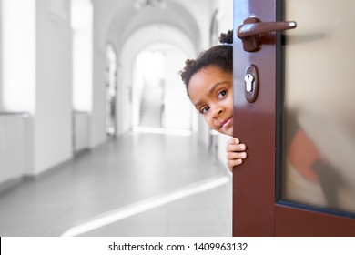 Pretty African Girl Hiding From Opened Door In School Corridor , Looking At Camera. Little And Cute Pupil Holding Door , Going To Lesson At Class Room. Concept Of Break.