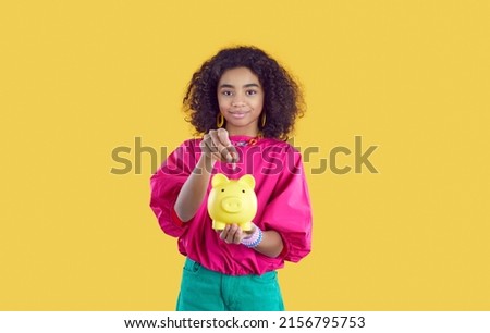 Pretty African girl holding piggybank isolated on yellow background. Child saving up money. Happy kid learning financial literacy. Studio shot of black school student putting coins inside piggy bank