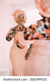 Pretty African Woman In Elegant Fashionable Pink Colorful Dress And Turban, Looking At Herself At Mirror, Admire Her Reflection