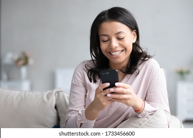 Pretty African young woman rest on sofa use smart phone smile look at screen chat with boyfriend read pleasant sms message media news on-line, modern tech every day usage for fun or business concept - Powered by Shutterstock