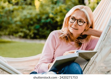 Pretty aged blonde woman in glasses reading a book literature novel in hammock in summer garden park outdoors, dreaming imagining - Powered by Shutterstock