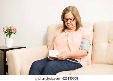 Pretty Aged Latin Woman Using Digital Pressure Cuff To Check Her Blood Pressure On A Couch At Home