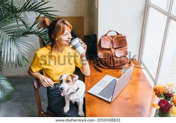Pretty\
amazing caucasian woman smile and looking aside with jack russel\
terrierwhile drinking coffee from reusable cup in loft cafe\
interior. Dog and people friendship\
concept.