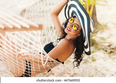Pretty amazing cheerful young girl on the beach, lying in a hammock and smiling in a black sexy bikini in a wide luxurious hat and mirrored sunglasses are reflected palm trees, lifestyle, tanned 