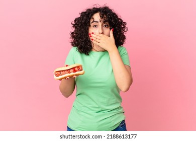 pretty arab woman covering mouth with hands with a shocked and holding a hot dog