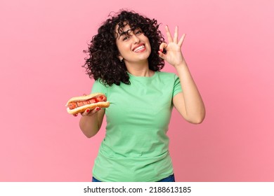 pretty arab woman feeling happy, showing approval with okay gesture and holding a hot dog