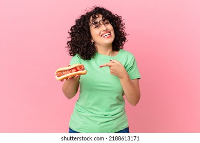 pretty arab woman looking excited and surprised pointing to the side and holding a hot dog