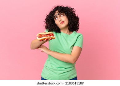 pretty arab woman shrugging, feeling confused and uncertain and holding a hot dog