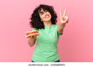 pretty arab woman smiling and looking happy, gesturing victory or peace and holding a hot dog