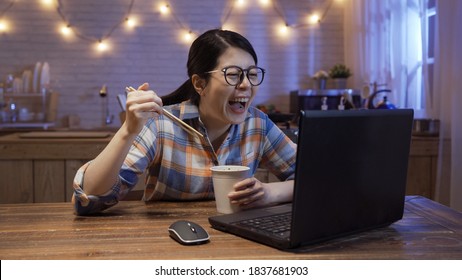 Pretty Asian Chinese Woman Hungrily Eating Ramen Soup And Joyfully Watching Series On Laptop Computer Late At Night In Kitchen.