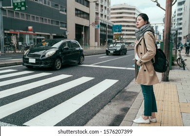 pretty asian chinese woman traveler walking in city osaka waiting for traffic lights to cross busy road with cars driving by. full length girl with bag standing on street and ready to pass pedestrian - Powered by Shutterstock