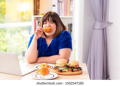 Pretty Asian fat woman eating fried chicken While working on the table Which has other food such as hamburgers and pizza, to food and health concept.