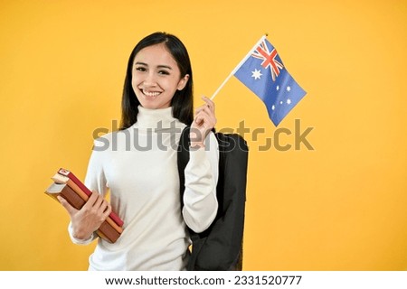 A pretty Asian female college student is holding her books and the Australia flag, standing against a yellow, isolated background. Exchange student and study abroad concept
