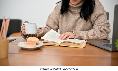 A Pretty Asian Female In Comfy Sweater Relaxes While Reading A Book And Sipping Her Hot Cocoa. Cropped Shot