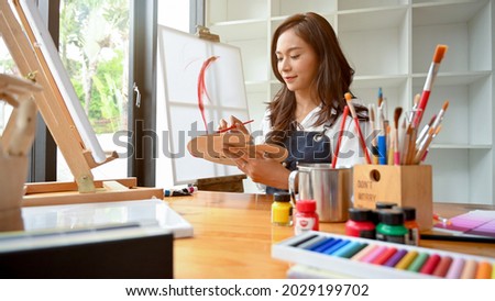 Pretty asian female painter do artwork in art workshop, painting supplies, oil pastels, two canvas easel, creative space