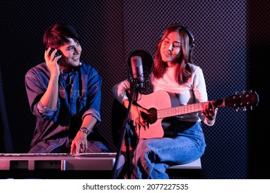 Pretty Asian female singer playing acoustic guitar with male playing electric keyboard. Recording songs by using a studio microphone and pop shield on microphone in blue and red light. Duet session.