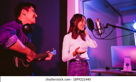 Pretty Asian female singer recording songs by using a studio microphone and pop shield on mic with male playing guitar in blue and red light. Performance and show in the music business. Duet session. - Powered by Shutterstock