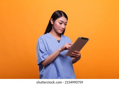 Pretty Asian Hospital Nurse Wearing Blue Medical Uniform While Using Modern Tablet On Orange Background. Clinic Healthcare Staff Member Using Digital Tablet. Studio Shoot.