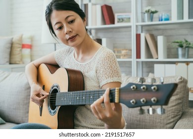 pretty asian Japanese lady in casual wear is plucking the string while learning to play a an acoustic guitar on the couch in a cozy bright living room at home.