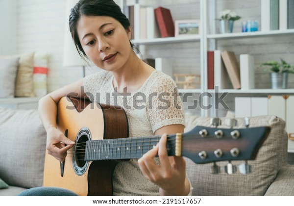 pretty asian Japanese lady in casual\
wear is plucking the string while learning to play a an acoustic\
guitar on the couch in a cozy bright living room at\
home.